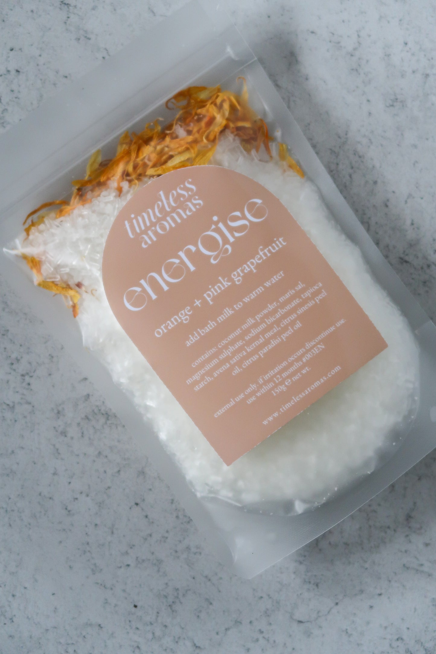 Timeless Aromas Energise Bath Milk displayed in a clear package, showcasing coconut milk and aromatic salts with floral decorations. Perfect for adding relaxation to your bath, this energise vegan bath milk is infused with citrus essential oils.