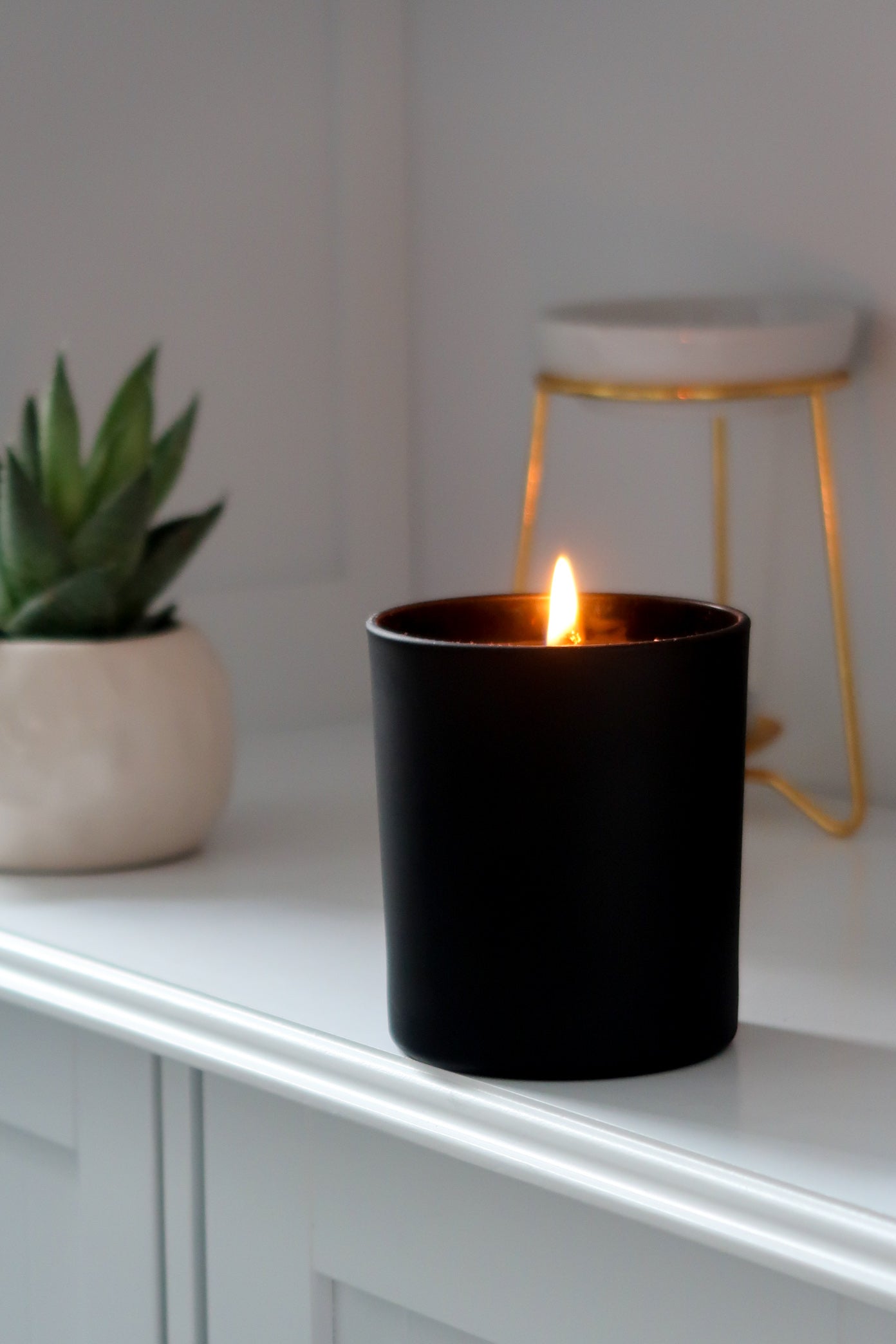 Another angle of the 30cl Black Matte Candle by Timeless Aromas Ltd, alight and glowing on a shelf. This stunning hand poured candle is housed in a ceramic jar, making it a wonderful choice for home fragrance. Its matte finish and blank label allow for custom branding, and it's sold in a pack of 6. This versatile candle works well as a gift or for wholesale opportunities, featuring a classic white candle inside.