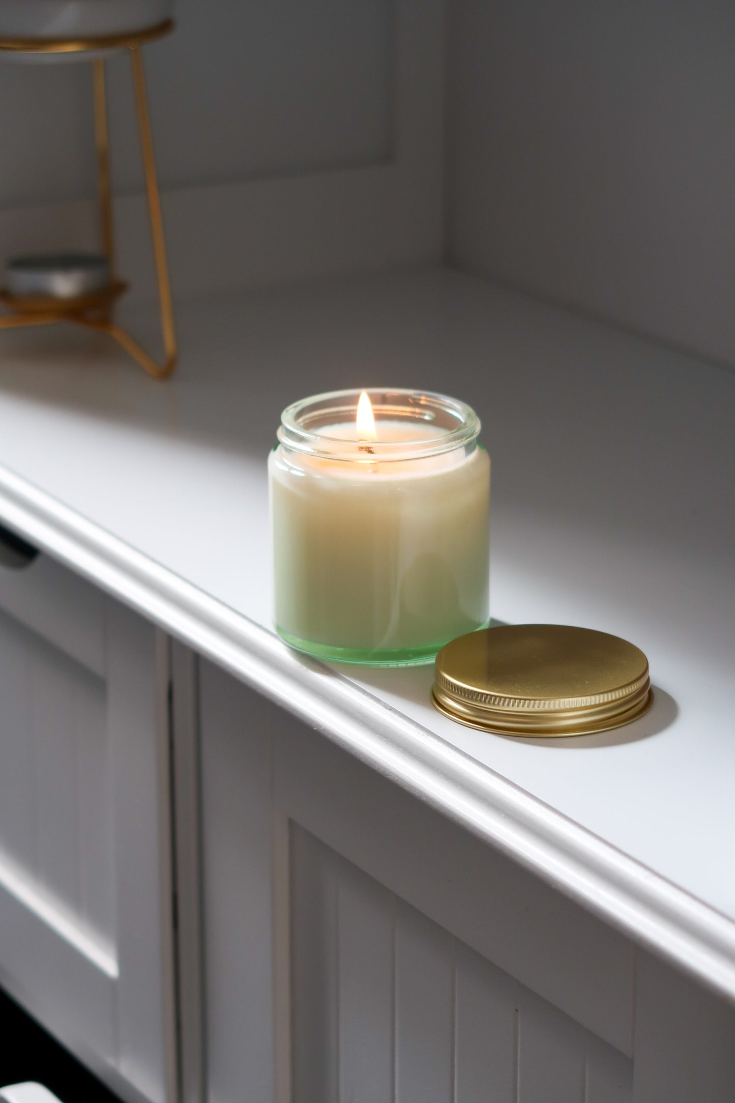 Another view of the 120g Clear Glass Gold Lid Candle from Timeless Aromas Ltd, showcasing the candle lit and glowing softly. This elegant hand poured candle is included in a pack of 6, featuring a classic gold lid, making it a great choice for home fragrance and gifts. Perfect for wholesale, it comes with a blank label.