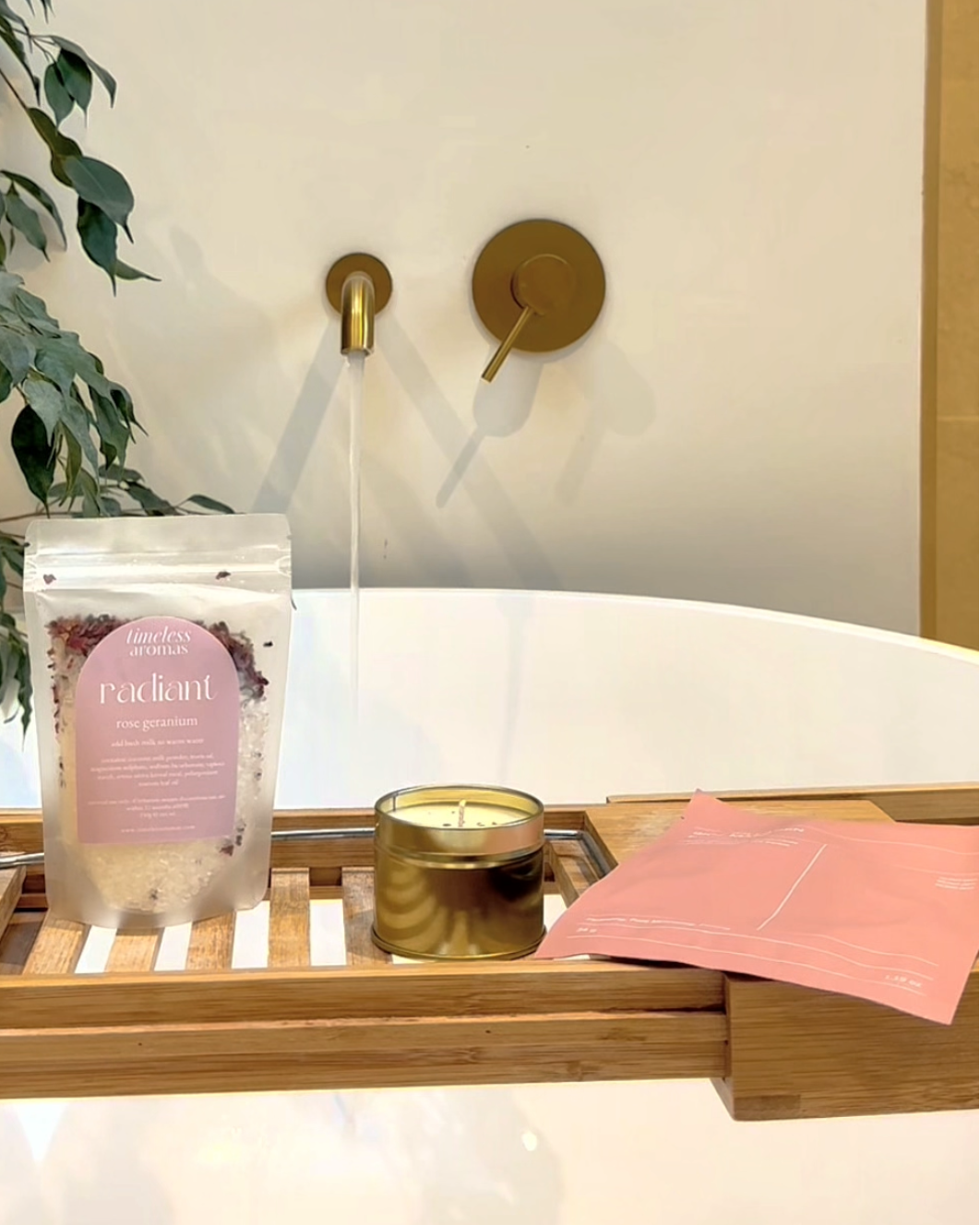 A bath scene with a wooden caddy holding a pink-labeled bath soak pouch, a gold candle tin, and a pink bath towel, set on a white bathtub with a brass faucet and plant in the background.