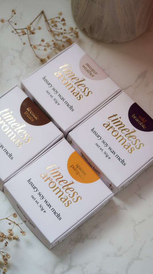 Four boxed scented wax melts from the brand Timeless Aromas arranged on a light marble surface with dried floral decor and a neutral vase in the background. The boxes are white with gold typography and colored circular labels in brown, orange, and dark purple, indicating different scent notes.