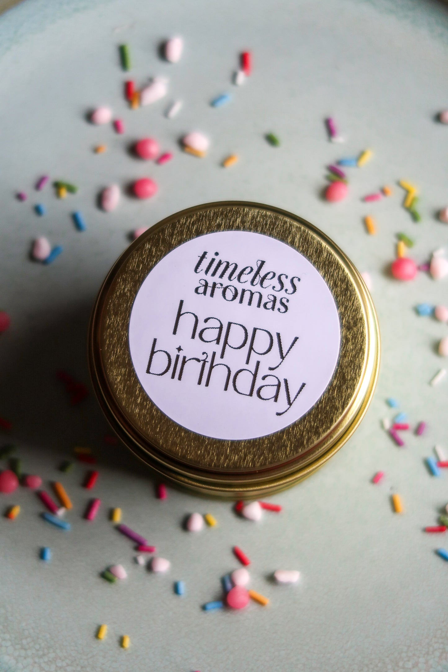 Stylish view of the Happy Birthday Travel Tin Candle from Timeless Aromas placed on a sprinkled plate, an excellent choice for a birthday tin candle gift.
