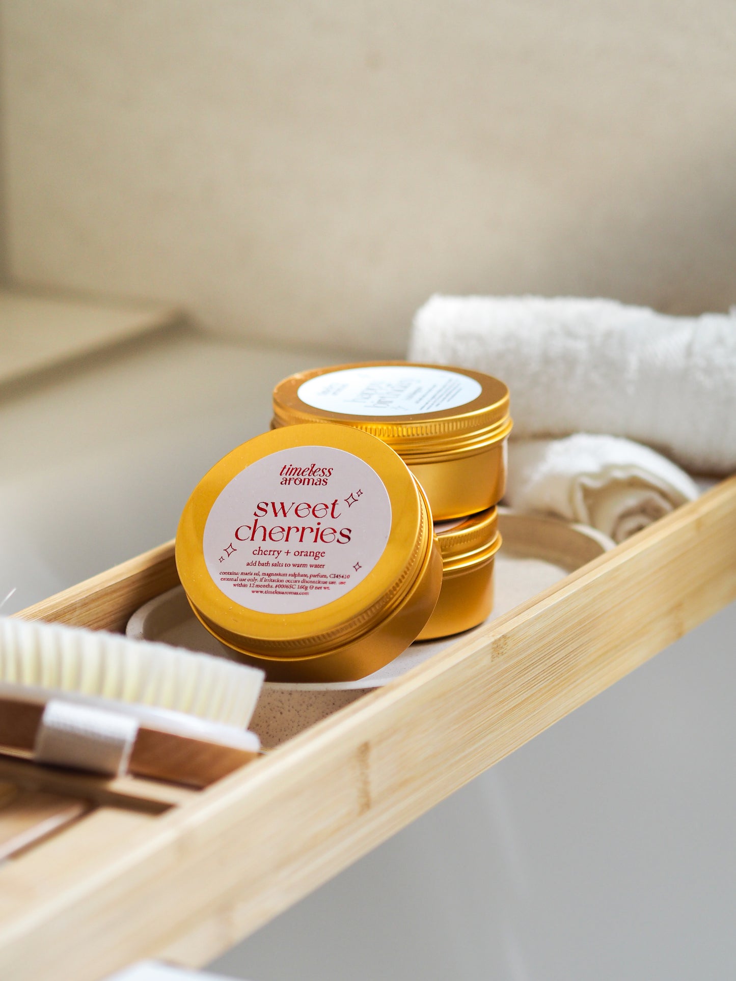 Timeless Aromas Sweet Cherries Gold Tin Bath Salts elegantly displayed with bath accessories, creating a serene spa-like atmosphere. A delightful option for bath salts for relaxation.