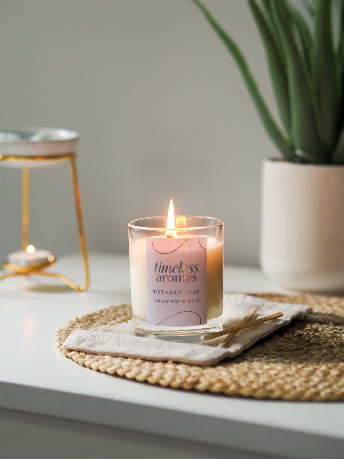 Timeless Aromas Birthday Cake Scented Candle burning warmly in a clear glass on a rustic placemat, perfect for a sweet home fragrance experience.