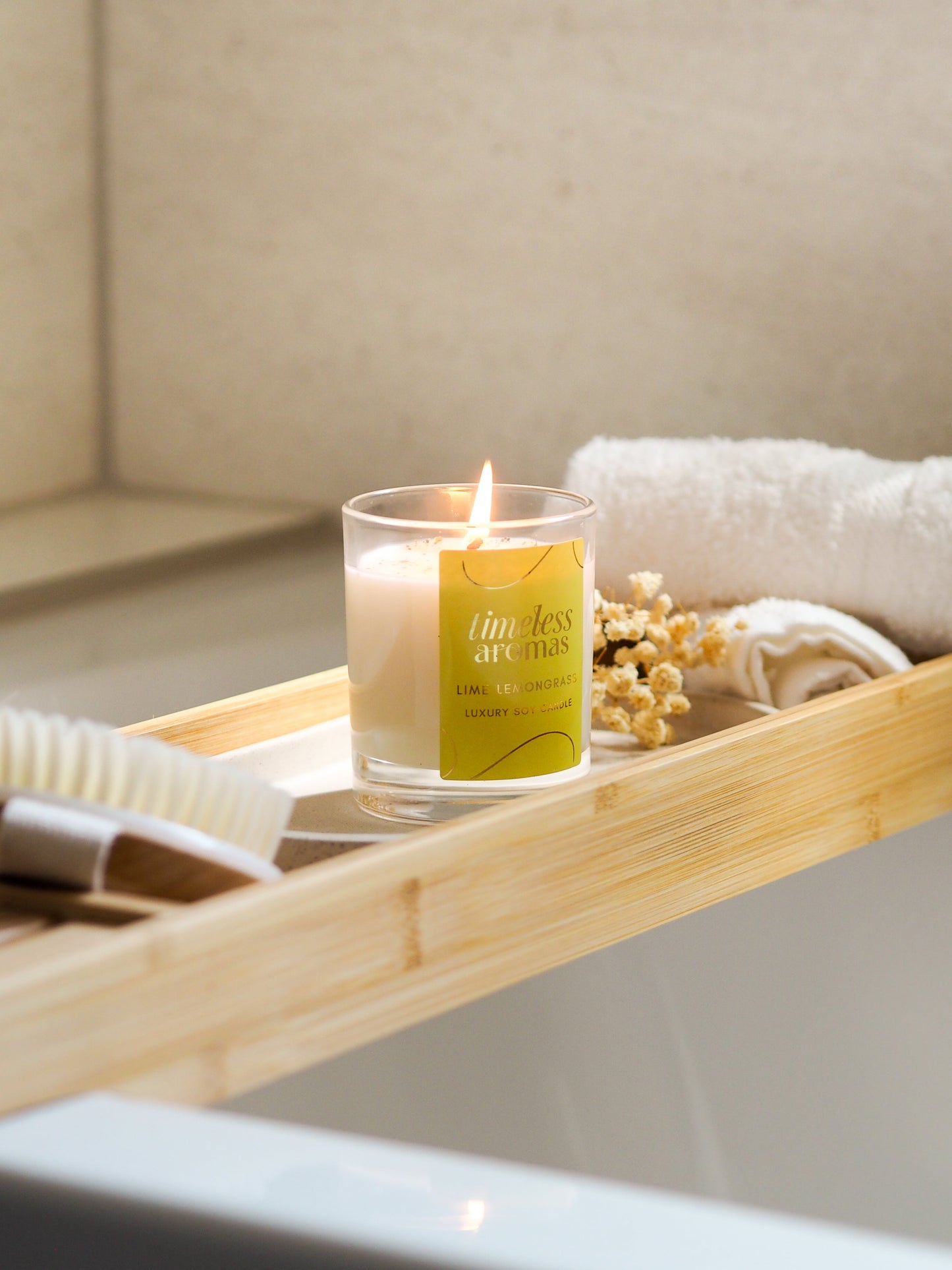 The beautifully lit Lime Lemongrass Luxury Candle by Timeless Aromas resting on a bamboo tray in a bathroom setting, complemented by towels and dried flowers.