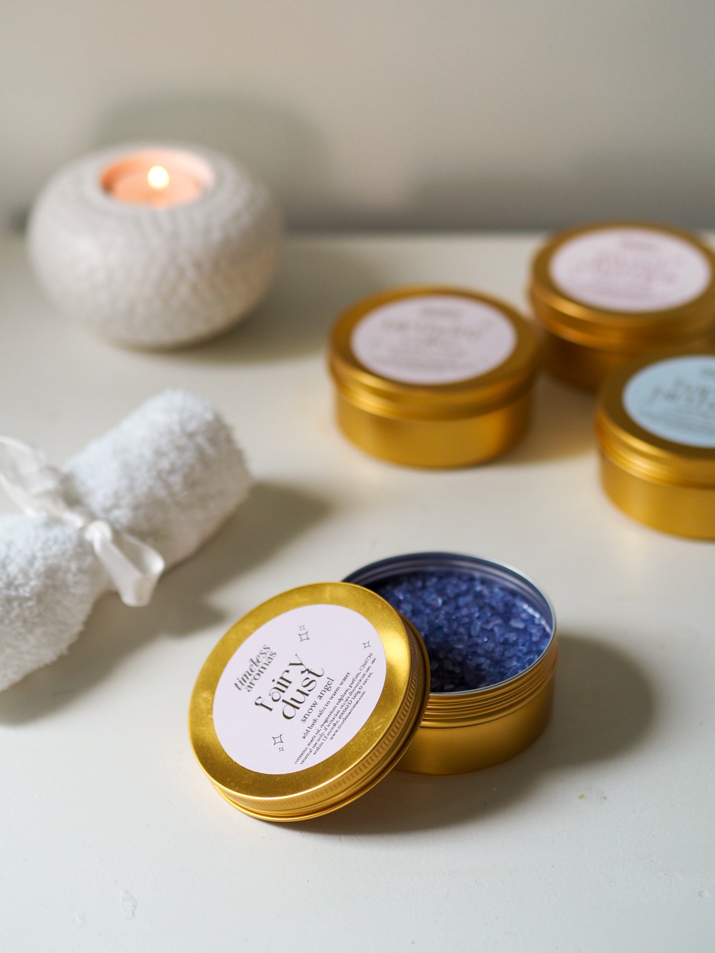 Open Fairy Dust Gold Tin Bath Salts by Timeless Aromas with vibrant purple salts, set elegantly next to a candle and a rolled towel, showcasing a perfect bath setup. These bath salts for relaxation create a calming atmosphere with their delightful Snow Angel fragrance.