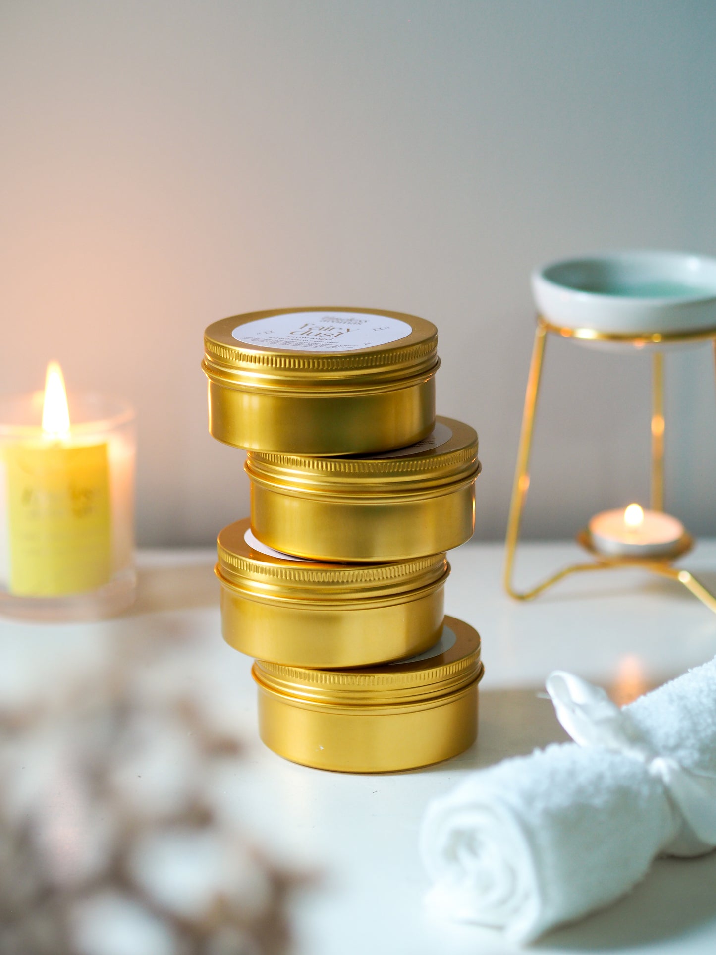 Four tins of Sweet Cherries Gold Tin Bath Salts by Timeless Aromas stacked with calming candles in the background, representing indulgent bath salts for relaxation.