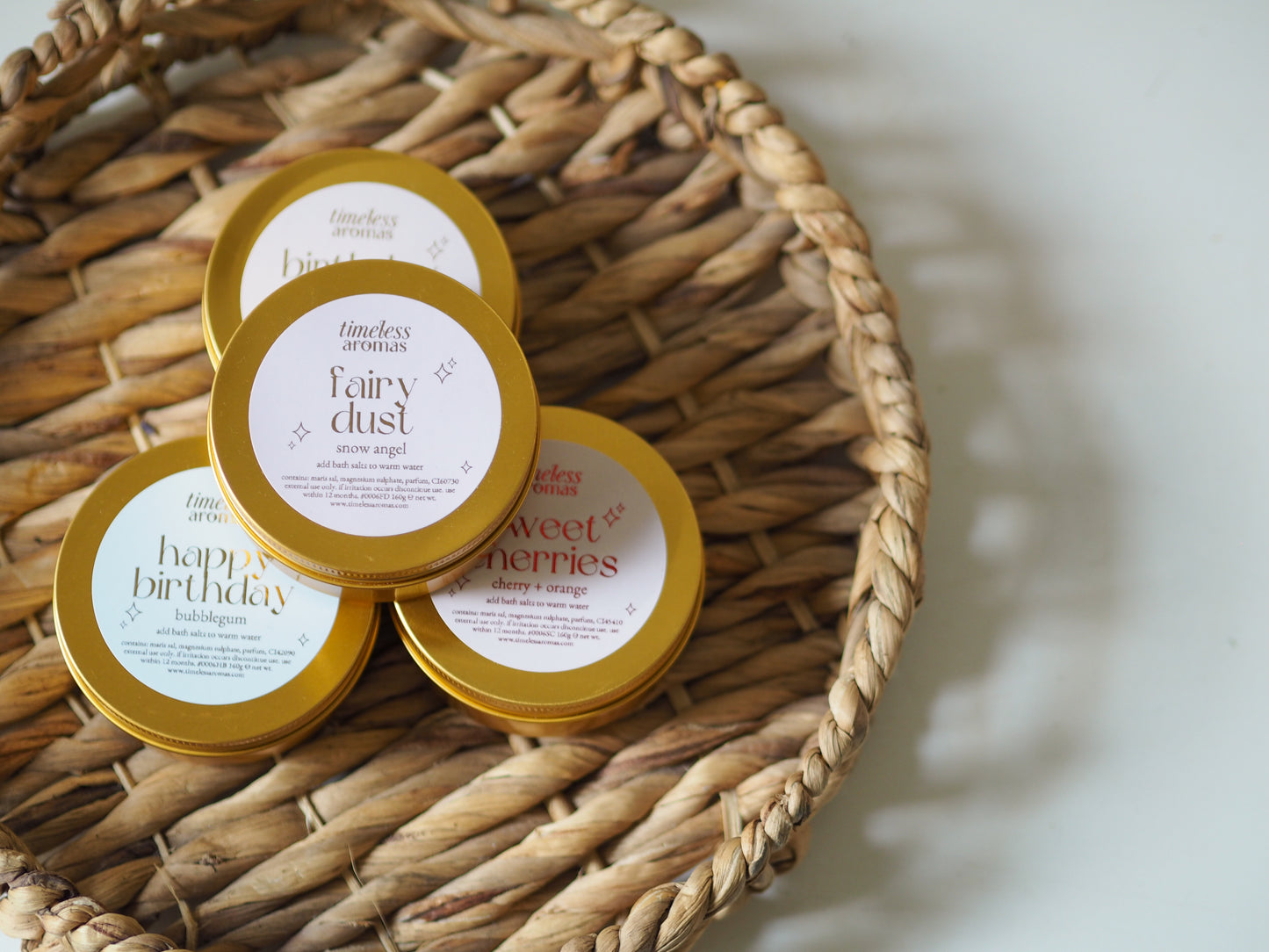 A collection of assorted bath salts including Sweet Cherries Gold Tin by Timeless Aromas, nestled in a woven basket, emphasising the luxury of bath salts for relaxation.
