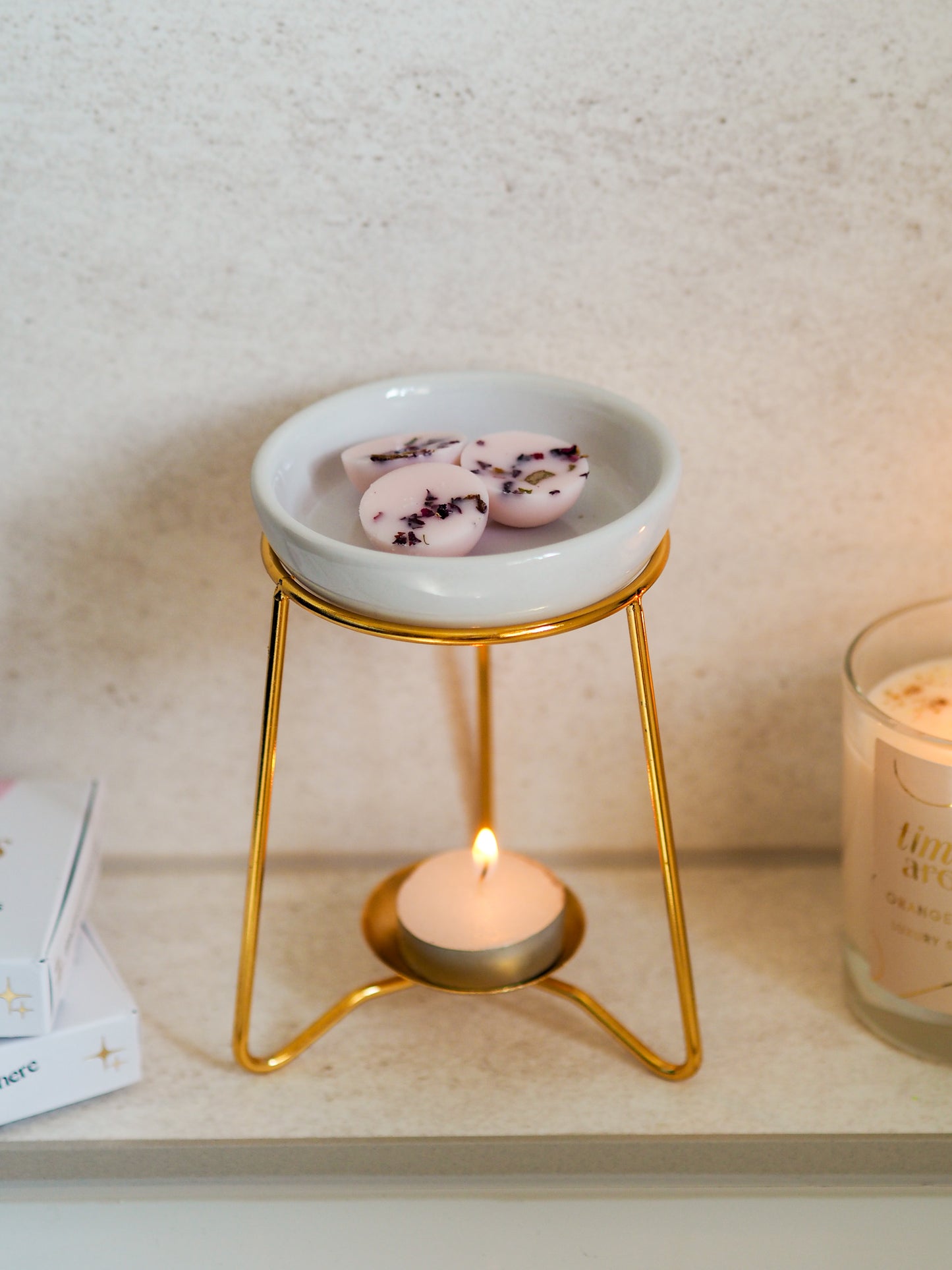 Close-up of a luxury wax melt burner featuring a gold frame and white ceramic dish, with lavender-scented wax melts melting above a tealight candle, from Timeless Aromas.