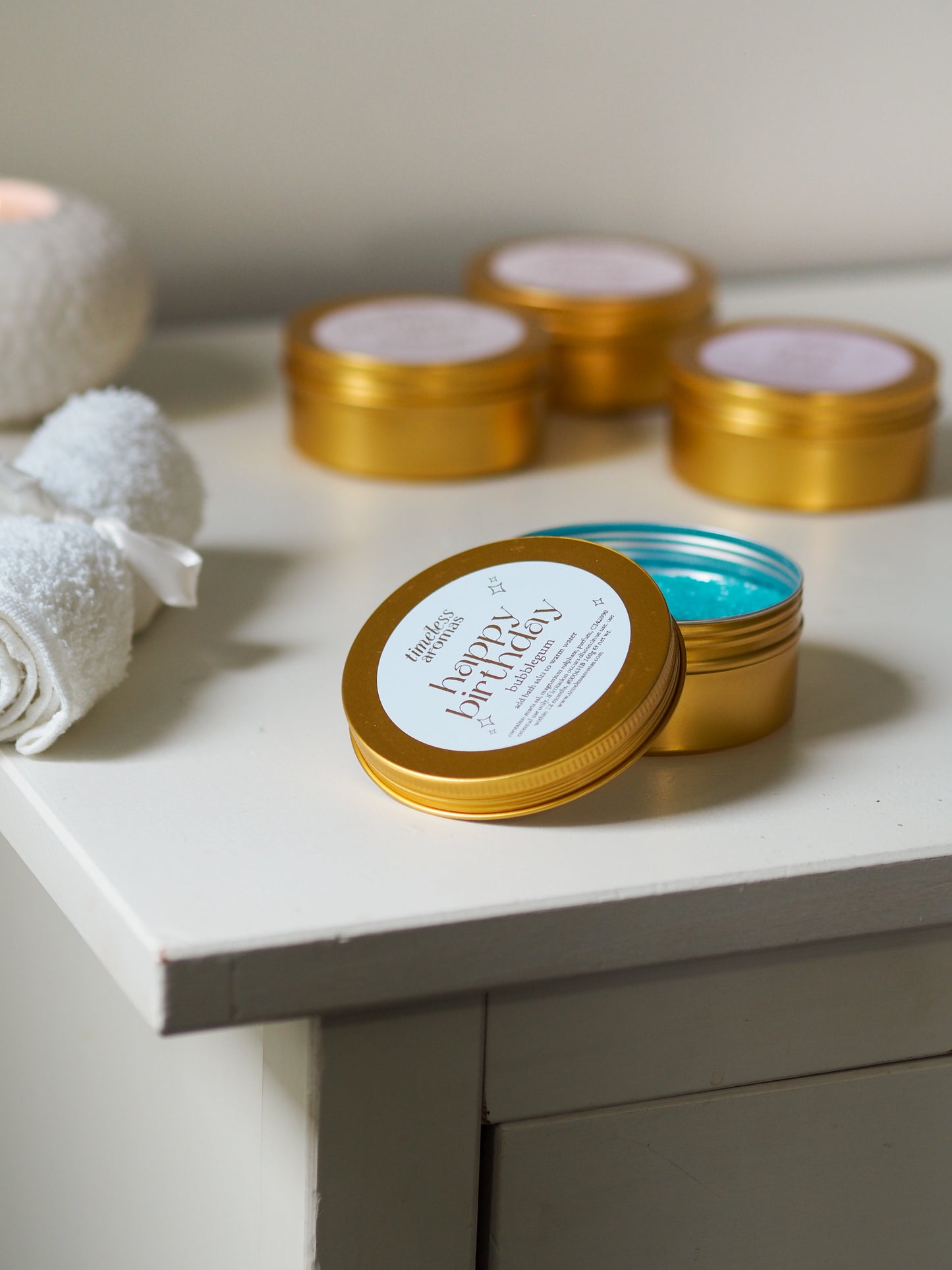 Open Happy Birthday Gold Tin Bath Salts by Timeless Aromas, showcasing blue bath salts ideal for relaxation, placed beside soft towels and lit candles.