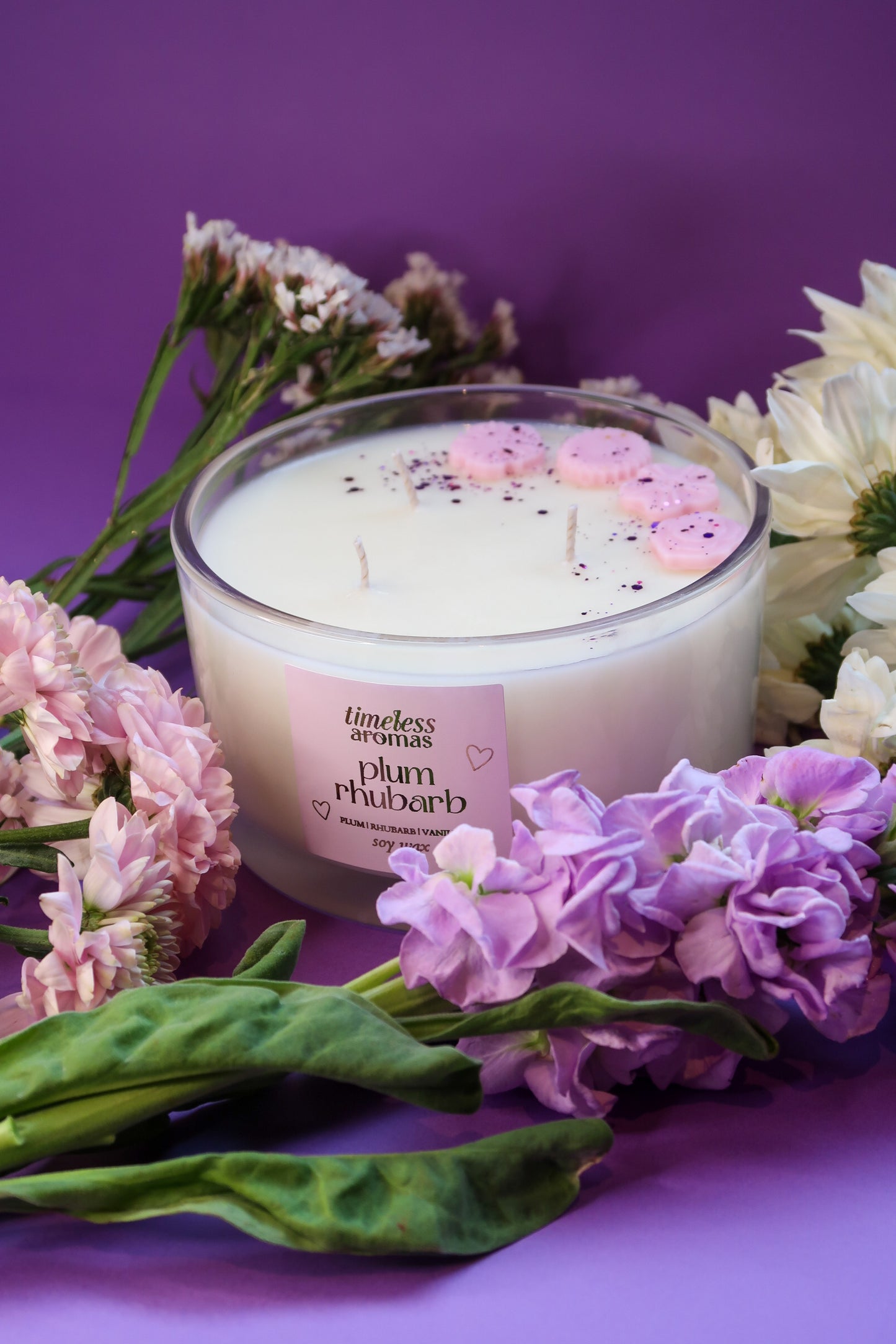 Timeless Aromas Plum Rhubarb Large Candle surrounded by flowers, showcasing its three wick design and decorative pink accents. Perfect for creating a cozy atmosphere with its fruity aroma.