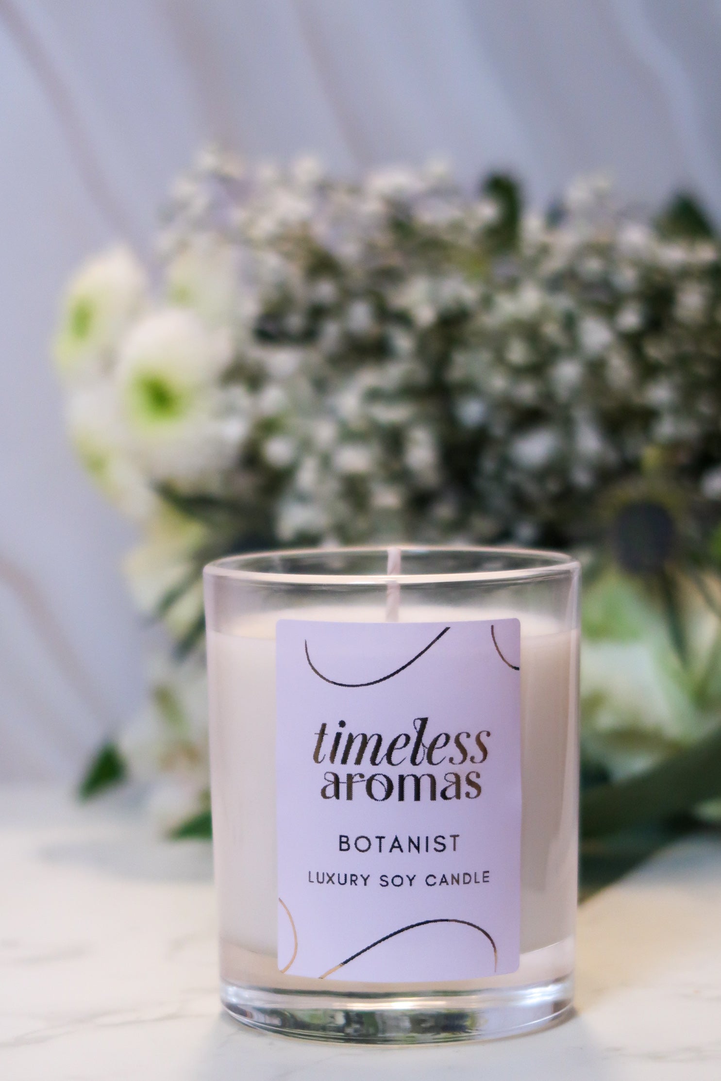 Timeless Aromas Botanist Handcrafted Luxury Candle displayed against a soft floral background, showcasing its elegant packaging.
