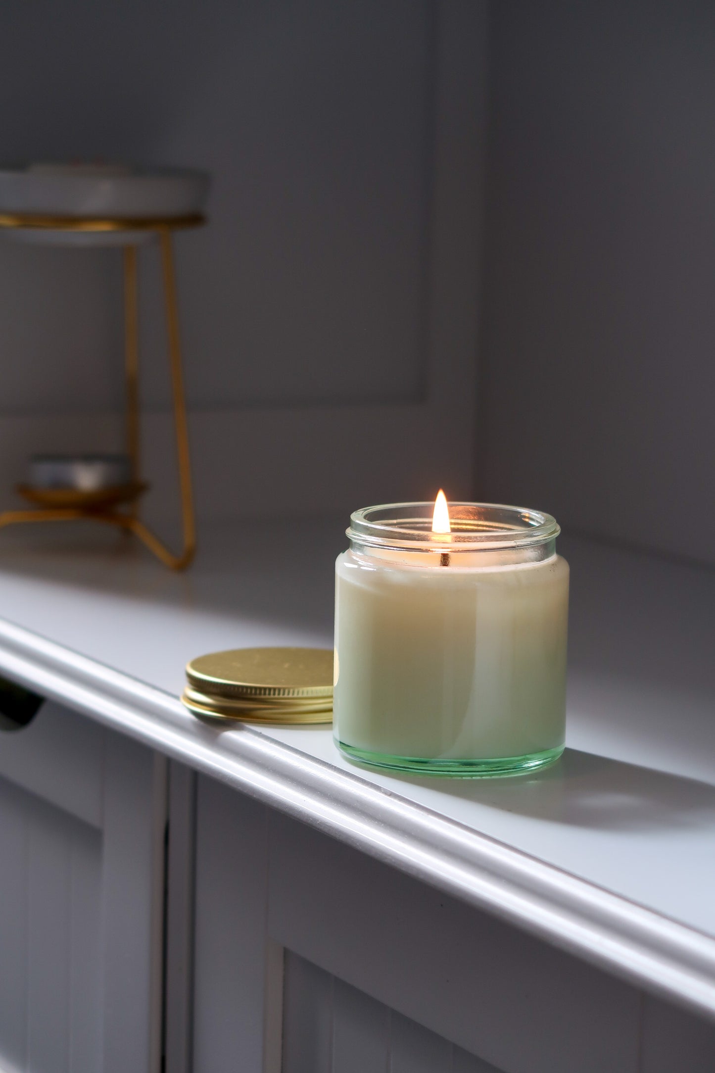 A lit 120g Clear Glass Gold Lid Candle from Timeless Aromas Ltd, part of a pack of 6 hand poured candles, showcasing its serene white wax. Ideal for home fragrance, this jar candle features a striking gold lid, perfect as a gift. Suitable for wholesale, it includes a blank label for customization.