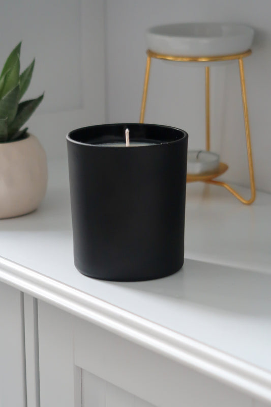 A 30cl Black Matte Candle from Timeless Aromas Ltd, displayed unlit on a shelf, highlighting its modern ceramic design. This hand poured candle is part of a pack of 6, perfect for home fragrance and gifting. The matte jar complements any decor and is suitable for wholesale options, featuring a blank label for personalisation. Ideal as a white candle in any space.