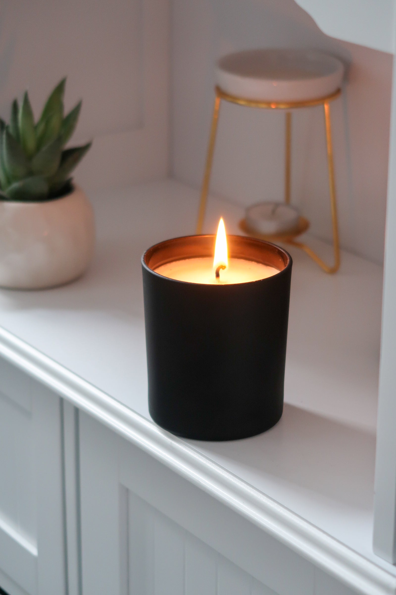 A lit 30cl Black Matte Candle by Timeless Aromas Ltd, radiating warmth from its flame. This hand poured candle stands out in its stylish ceramic jar, making it a chic addition to home fragrance collections. With a matte finish and blank label, this candle is part of a pack of 6, perfect for gifting or wholesale. The jar candle serves as an elegant white candle that fits any setting.