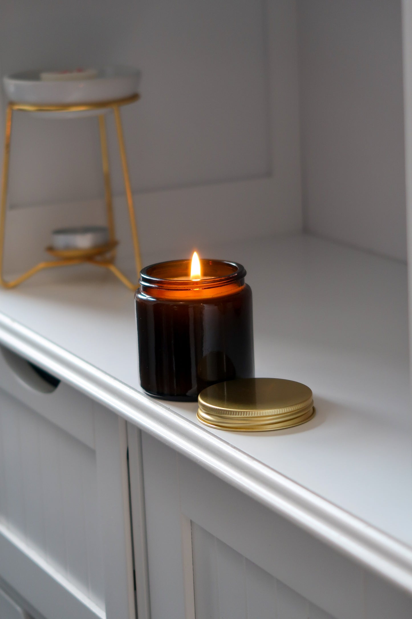 A lit amber glass jar candle with a gold lid from Timeless Aromas Ltd, included in the Amber Glass Candles 120g - Pack of 6. These hand poured candles are 12cl each, perfect for gifting or enhancing home fragrance as a stylish addition or for wholesale, and feature a blank label.