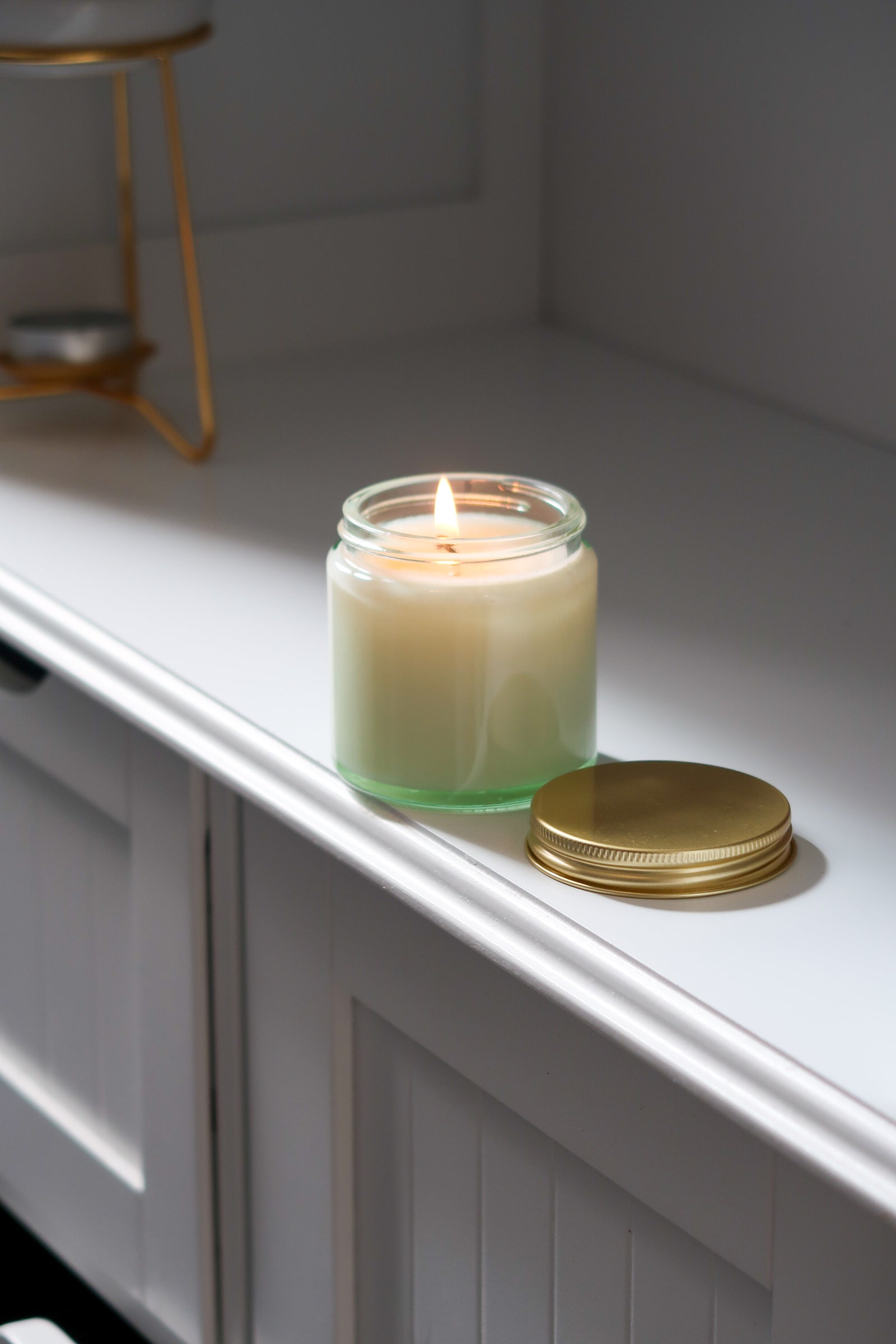 Another view of the 120g Clear Glass Gold Lid Candle from Timeless Aromas Ltd, showcasing the candle lit and glowing softly. This elegant hand poured candle is included in a pack of 6, featuring a classic gold lid, making it a great choice for home fragrance and gifts. Perfect for wholesale, it comes with a blank label.
