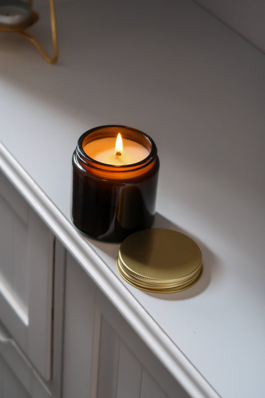 Hand poured candles in amber glass jars with gold lids from Timeless Aromas Ltd, featured as part of the Amber Glass Candles 120g - Pack of 6. These decorative 12cl jar candles are perfect for home fragrance, making an elegant gift, or for wholesale. Each candle comes with a blank label, suitable for any occasion.