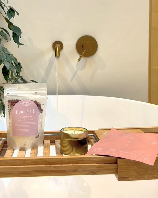 A bath scene with a wooden caddy holding a pink-labeled bath soak pouch, a gold candle tin, and a pink bath towel, set on a white bathtub with a brass faucet and plant in the background.