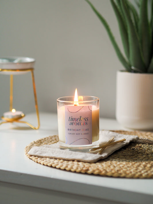 Timeless Aromas Birthday Cake Scented Candle burning warmly in a clear glass on a rustic placemat, perfect for a sweet home fragrance experience.