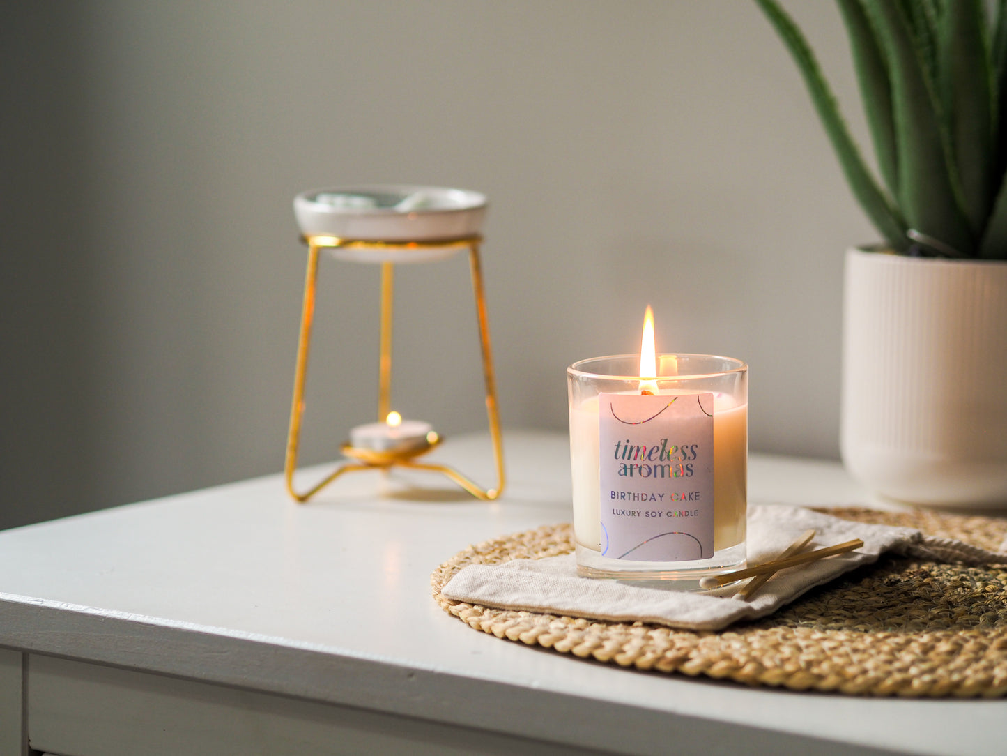 Timeless Aromas Birthday Cake Scented Candle glowing brightly on a table beside a golden tea light holder, adding a festive touch to home decor.