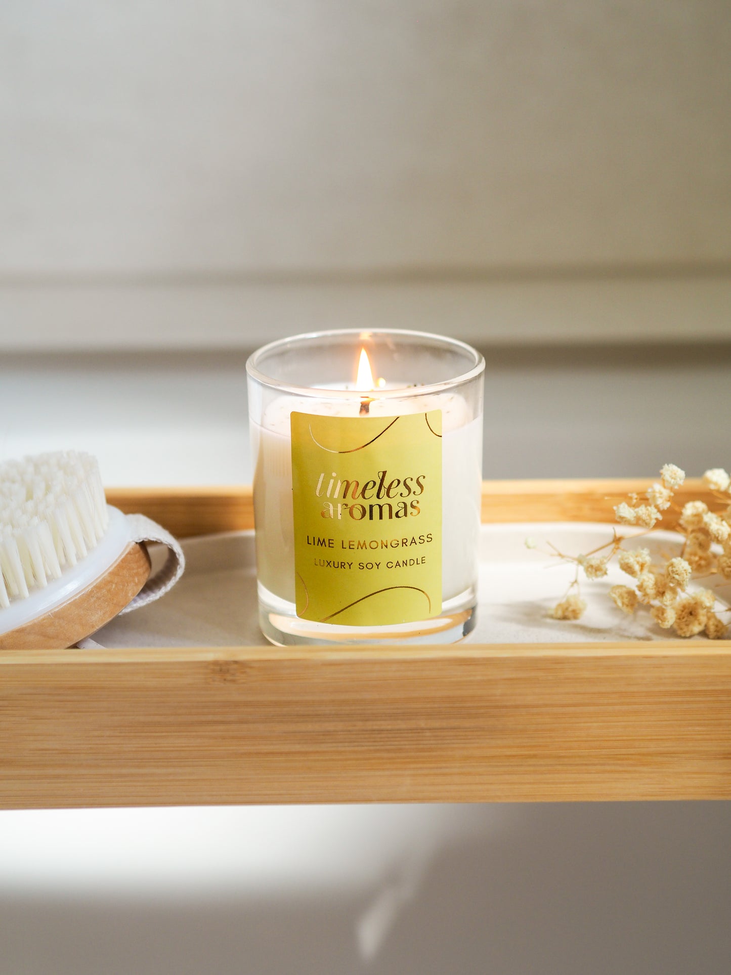 A lit Lime Lemongrass Luxury Candle by Timeless Aromas on a bamboo tray, surrounded by a natural brush and dried flowers, enhancing the refreshing ambiance.