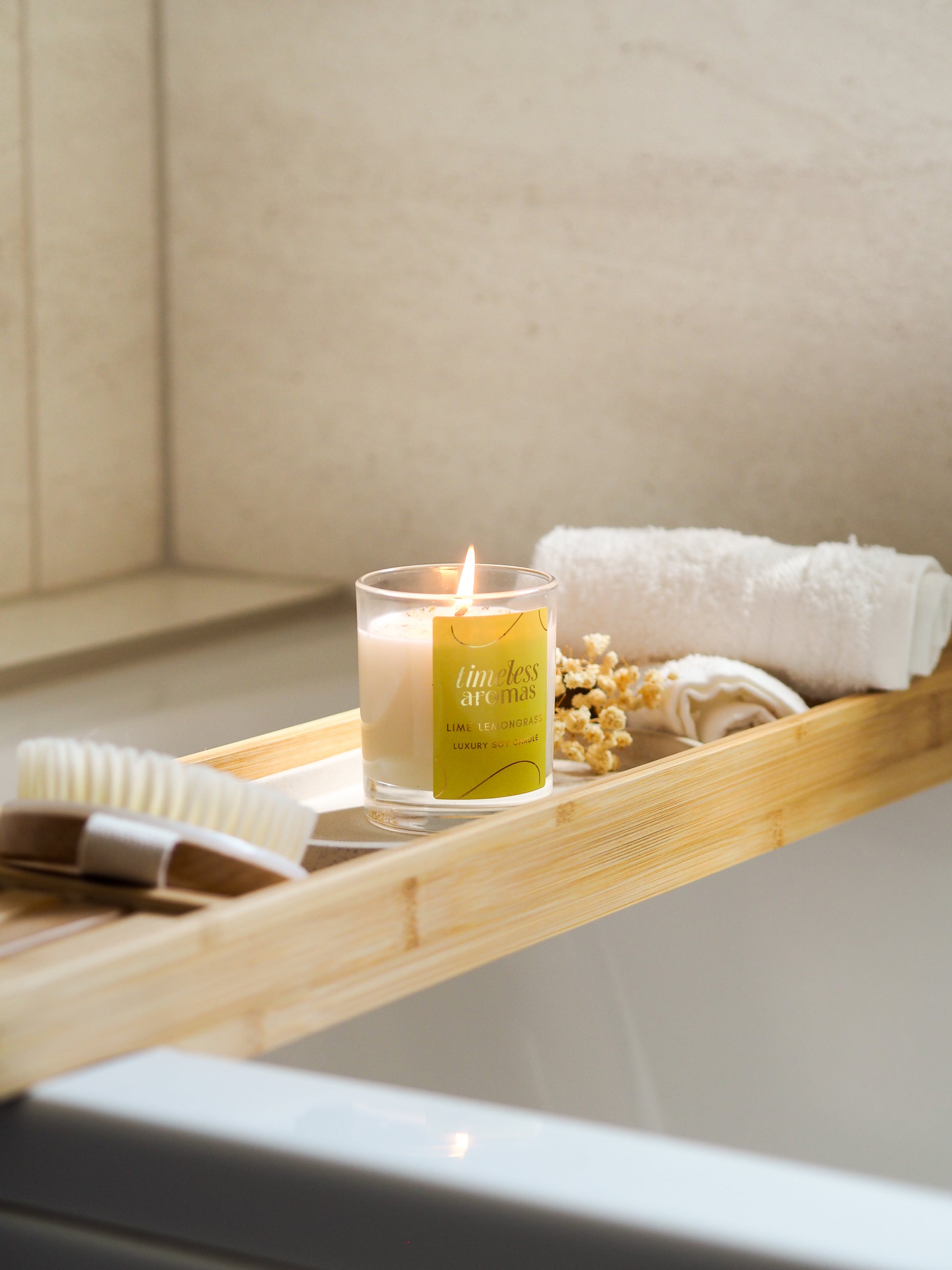 A glowing Lime Lemongrass Luxury Candle by Timeless Aromas placed on a bamboo bath tray beside towels and decorative flowers, creating a tranquil setting.