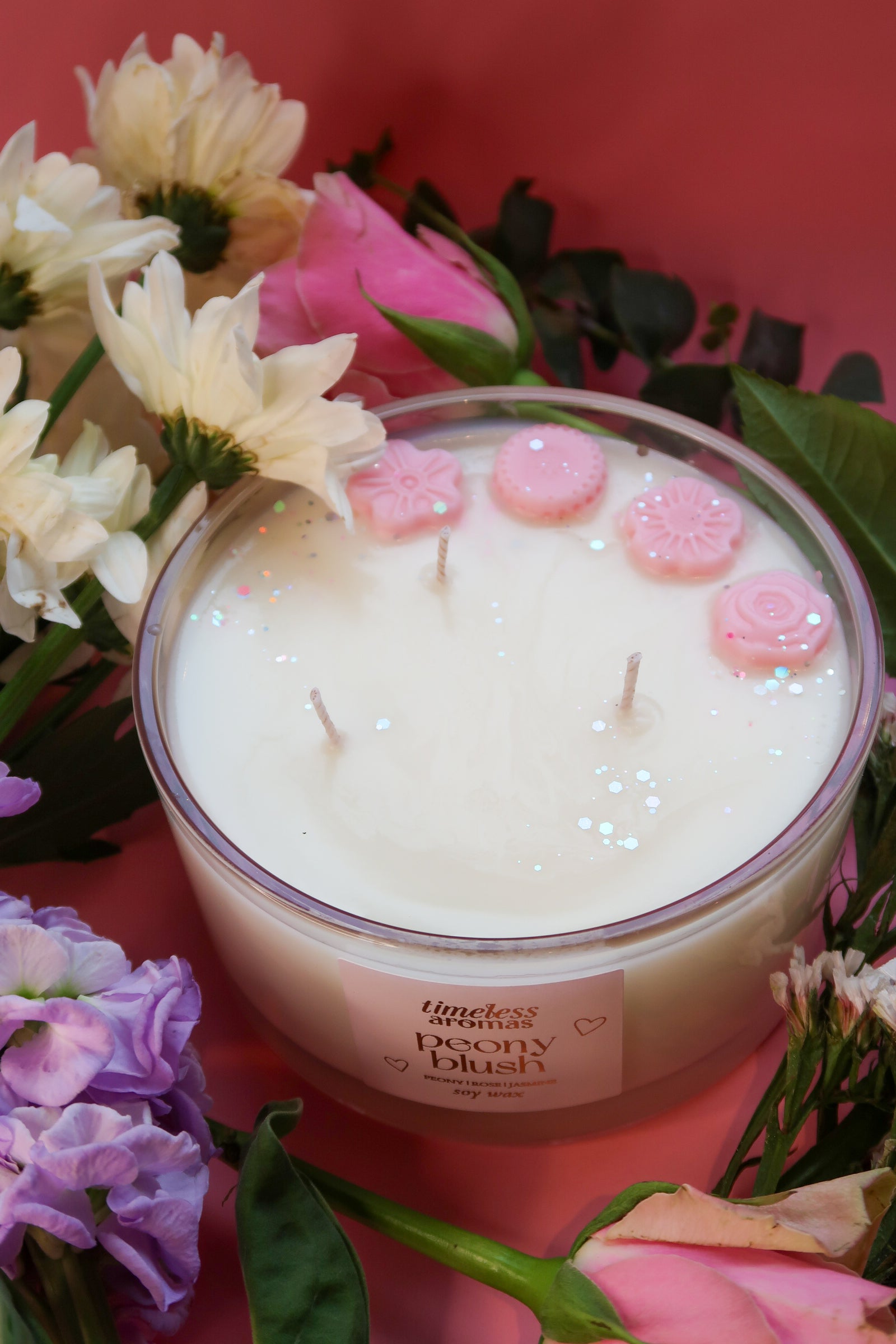 Close-up of Timeless Aromas Peony Blush Large Candle showcasing three wicks, topped with decorative pink elements, nestled among fresh flowers, representing elegance and charm.