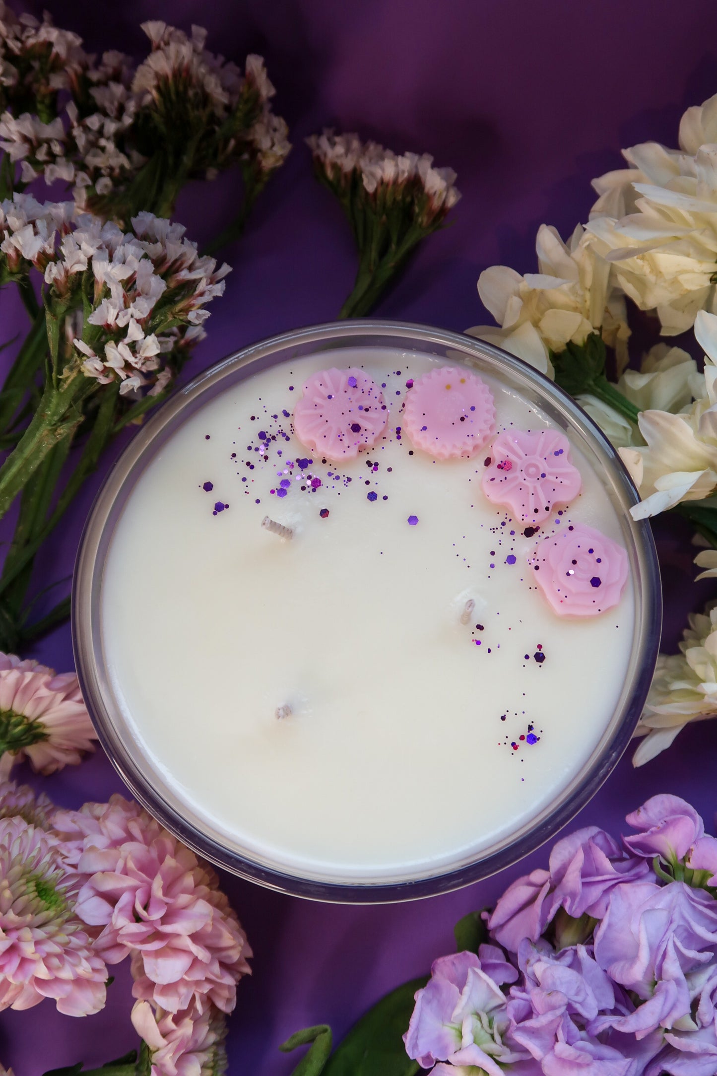 Top view of Timeless Aromas Plum Rhubarb Large Candle, featuring a three wick design adorned with pink decorations and surrounded by vibrant flowers on a purple background, ideal for gifting or home decor.